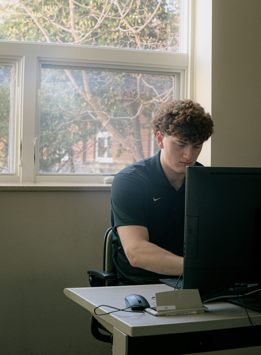 A student with curly hair sits at a desk working on a desktop computer in a brightly lit room. Sunlight streams through a large window behind him, revealing leafless tree branches and a brick building outside. He is wearing a dark Nike polo shirt and appears focused on the screen. A mouse, keyboard, and small desk placard labeled "41" are visible on the desk.